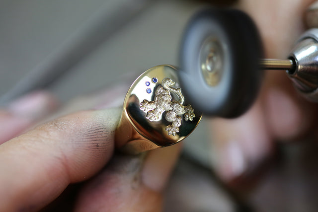 Gold ring with intricate design and three blue sapphire stones held by a hand, with a tool in the background.